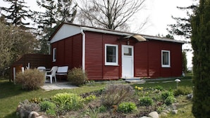A red wooden cabin with a white door and windows, surrounded by a garden with various plants and rocks.