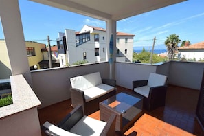 Un balcón con mobiliario de mimbre y una mesa de vidrio, con vistas a una zona residencial con edificios y un cielo despejado.