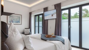 A modern hotel room with a large bed, a flat-screen TV, a dining table with breakfast, and a sliding glass door to the outside. - Cozy apartment in Binz with outdoor swimming pool (Prora/Rügen)