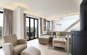 Un salon moderne avec un canapé, des fauteuils et une table basse. Il y a une grande fenêtre avec des rideaux, une cheminée et une vue sur l’extérieur.