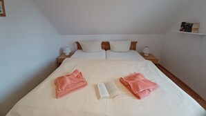 A neatly made bed with two pillows, a book, and two folded towels. There are two bedside tables with lamps, and a shelf with books on the wall. - By the Sea, Port Side - By the Sea - Port Side (Wustrow)