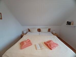 A neatly made bed with two pillows, a book, and two folded towels. There are two bedside tables with lamps, and a shelf with books on the wall. - By the Sea, Port Side - By the Sea - Port Side (Wustrow)