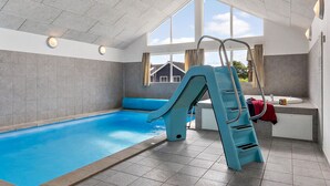 An indoor swimming pool with a blue slide, a ladder, and a view of a house through large windows. - Nice home in Sydals with sauna (Sydals)