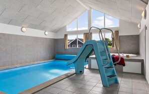 An indoor swimming pool with a blue slide, a ladder, and a view of a house through large windows.