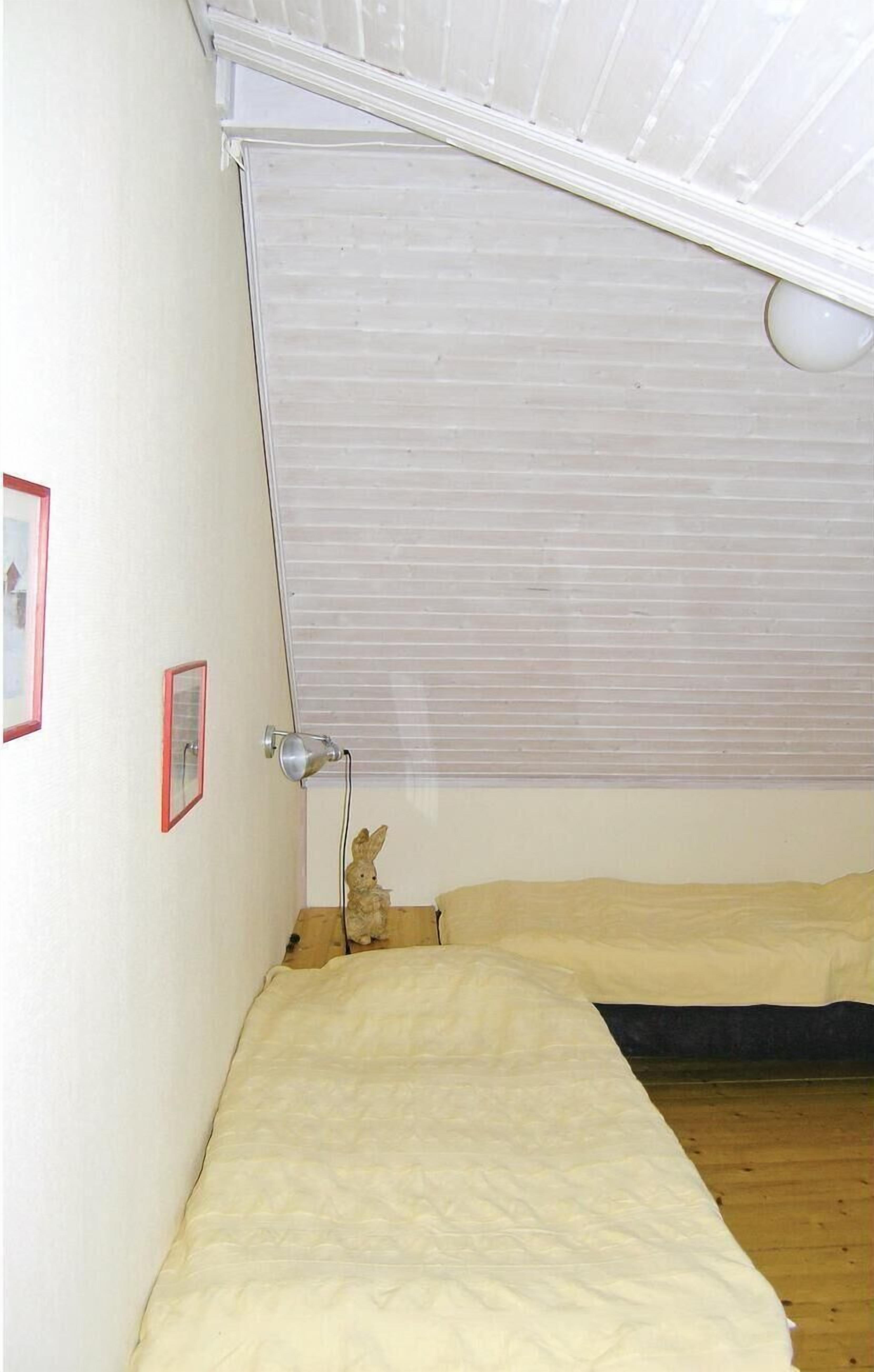 Una habitación pequeña con cama individual, techo inclinado, ventana con persianas, una lámpara de noche y un peluche pequeño sobre la cama.