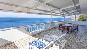 Un'area esterna coperta con vista sul mare, pavimento in pietra, sedie bianche e un tavolo.