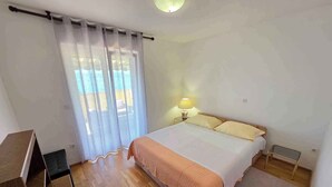 A bedroom with a bed, a nightstand with a lamp, a wooden desk, and a window with sheer curtains.