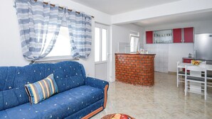 A living room with a blue sofa, a small table with a fruit bowl, and a kitchen area with a brick oven and red cabinets. - Beach front home in Osobjava with house sea view (Osobjava)