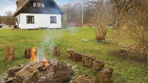 A white house with a black roof, a fire pit in the foreground, and a grassy yard with trees. - Amazing home in Askeby with kitchen (Askeby)