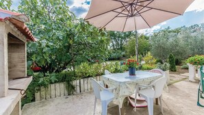 Een terras buiten met een tafel, stoelen, een grote parasol en verschillende planten en bomen.