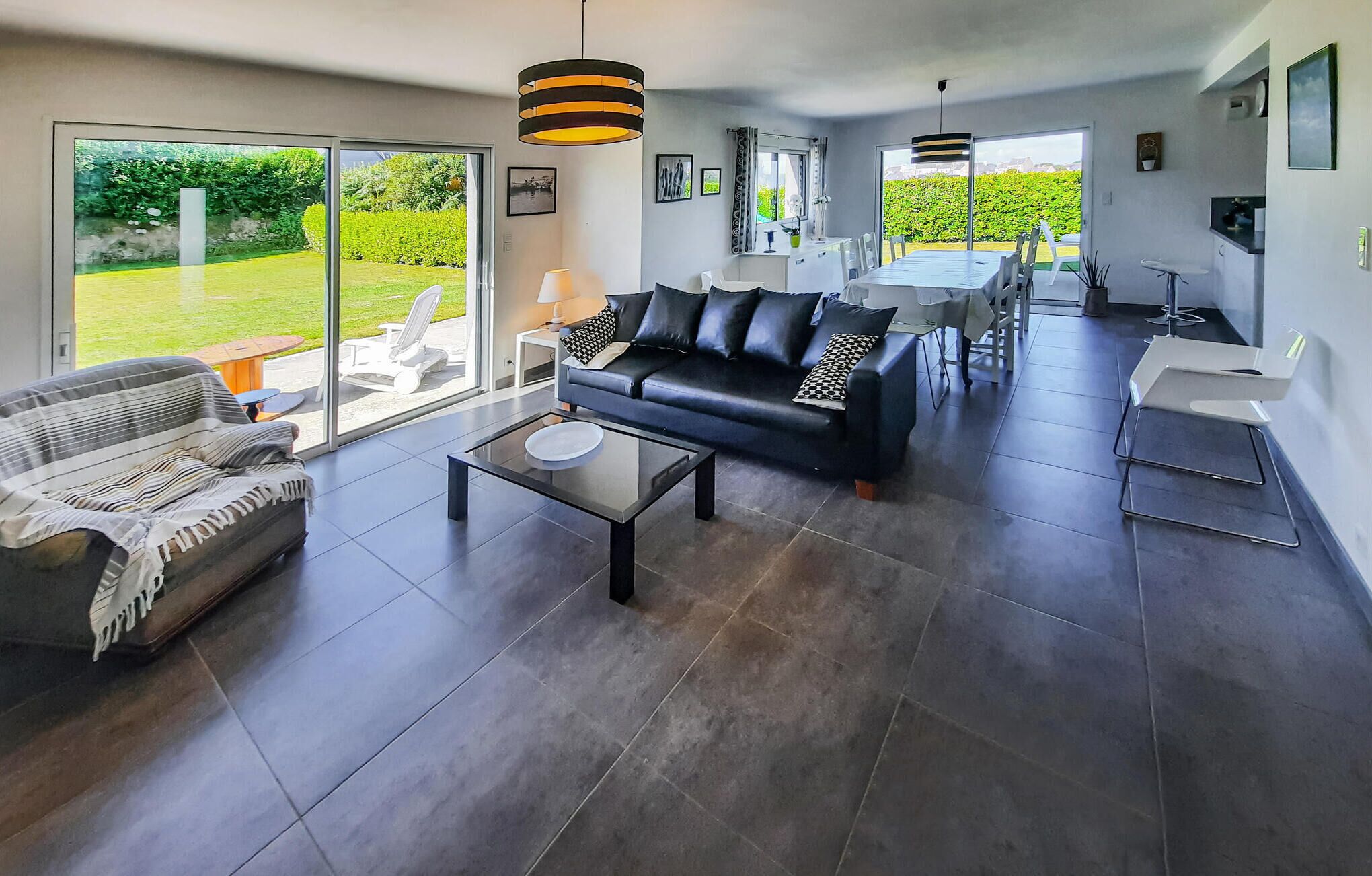 Ein großzügiger Wohnbereich mit einer schwarzen Ledersofa, einem gestreiften Sofa und einem Glastisch. Es gibt einen Essbereich mit Tisch und Stühlen, und große Fenster mit Blick auf den Garten.