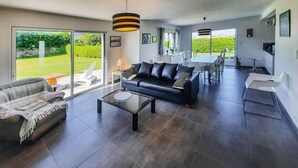 Een ruime zithoek met een zwarte leren bank, een gestreepte bank en een glazen salontafel. Er is een eethoek met een tafel en stoelen, en grote ramen met uitzicht op de tuin.