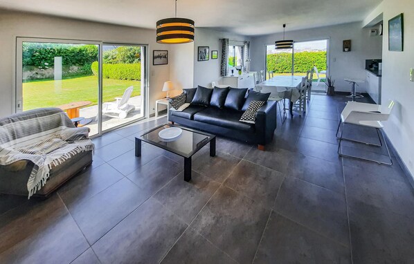 A spacious living area with a black leather sofa, a striped sofa, and a glass coffee table. There is a dining area with a table and chairs, and large windows offering a view of the garden. - Lovely home in Plouguerneau with WiFi (Plouguerneau)