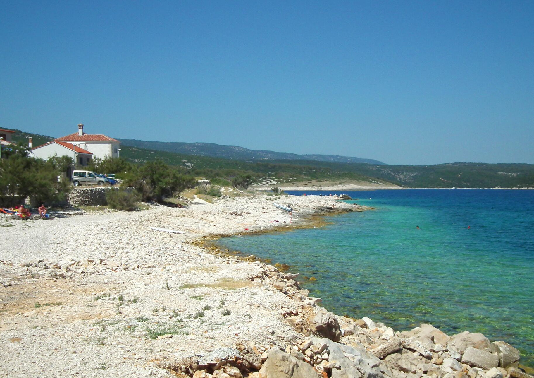 Eine Küstenlandschaft mit Kiesstrand, klarem blauen Wasser und einem Gebäude mit rotem Dach.