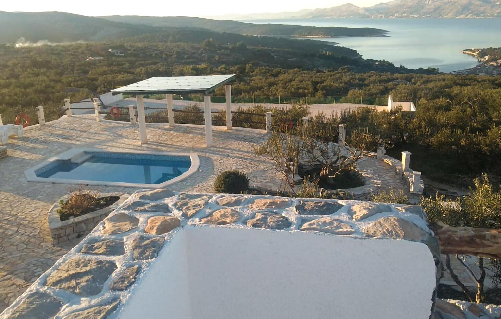 A stone pool with a pergola, surrounded by stone walls and vegetation, overlooking a coastal landscape.