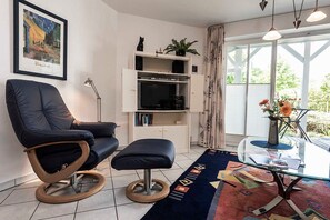 A modern living room with a black leather chair, a glass coffee table, a bookshelf, and a large window with curtains. - Am Stadtwald Apartment 3 - Am Stadtwald Strandstr. 15, WE 3 (Kühlungsborn)