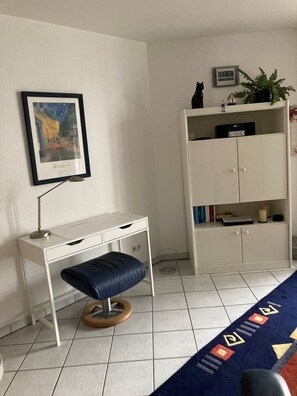 Une pièce avec un bureau blanc, un tabouret bleu rembourré, une armoire blanche avec des étagères, un tableau encadré au mur et une horloge.