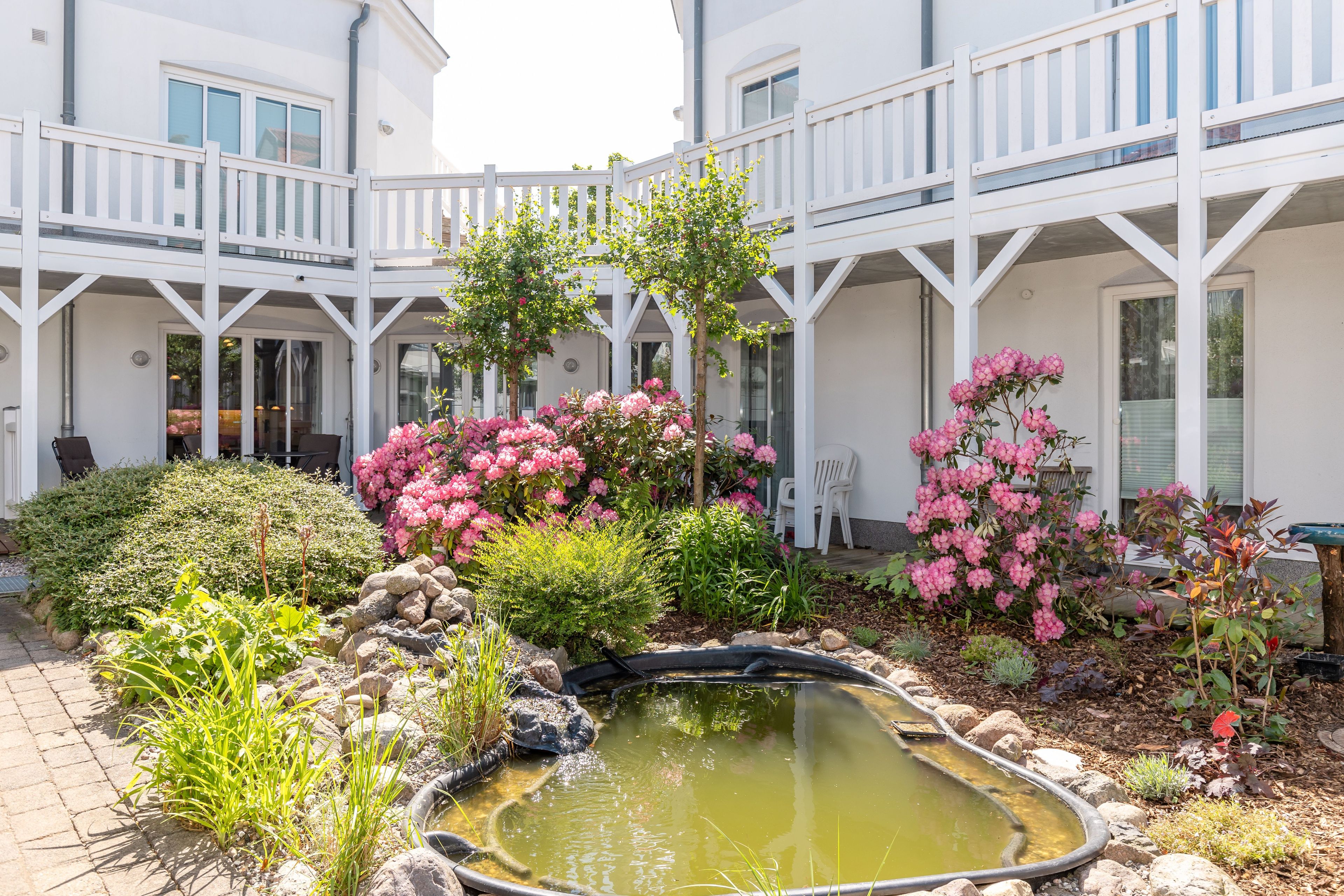 Ein Innenhof mit einem kleinen Teich, umgeben von weißen Geländern, rosa Blüten und verschiedenen Pflanzen.