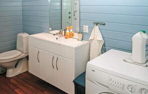 Une salle de bain avec un lavabo, des toilettes et un lave-linge, tous de couleur blanche. On trouve des articles de toilette sur le lavabo et une serviette est accrochée au mur.