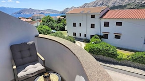 Un balcon avec un fauteuil rembourré et une petite table, donnant sur une ville côtière aux bâtiments blancs et offrant une vue sur la mer.
