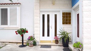 Ein Gebäudeeingang mit einer weißen Tür, einem kleinen Fenster mit Fensterläden und Topfpflanzen.