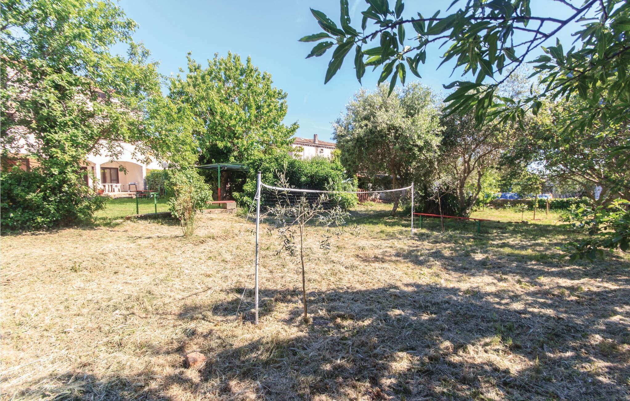 A fenced outdoor area with a volleyball net, surrounded by trees and a building in the background.
