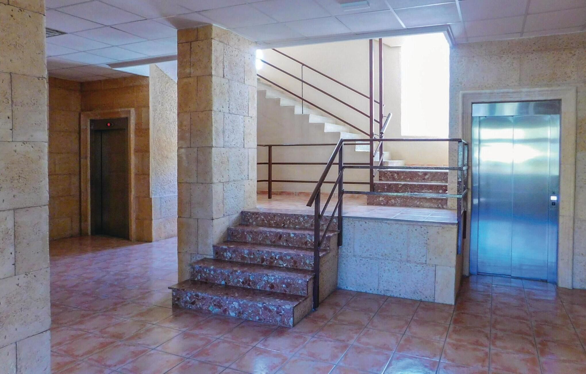A staircase with metal railings leading to an elevator, surrounded by stone pillars and tiled flooring.