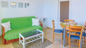 Een kleine, lichte kamer met een groene bank, een klein tafeltje met stoelen, en een witte kast.