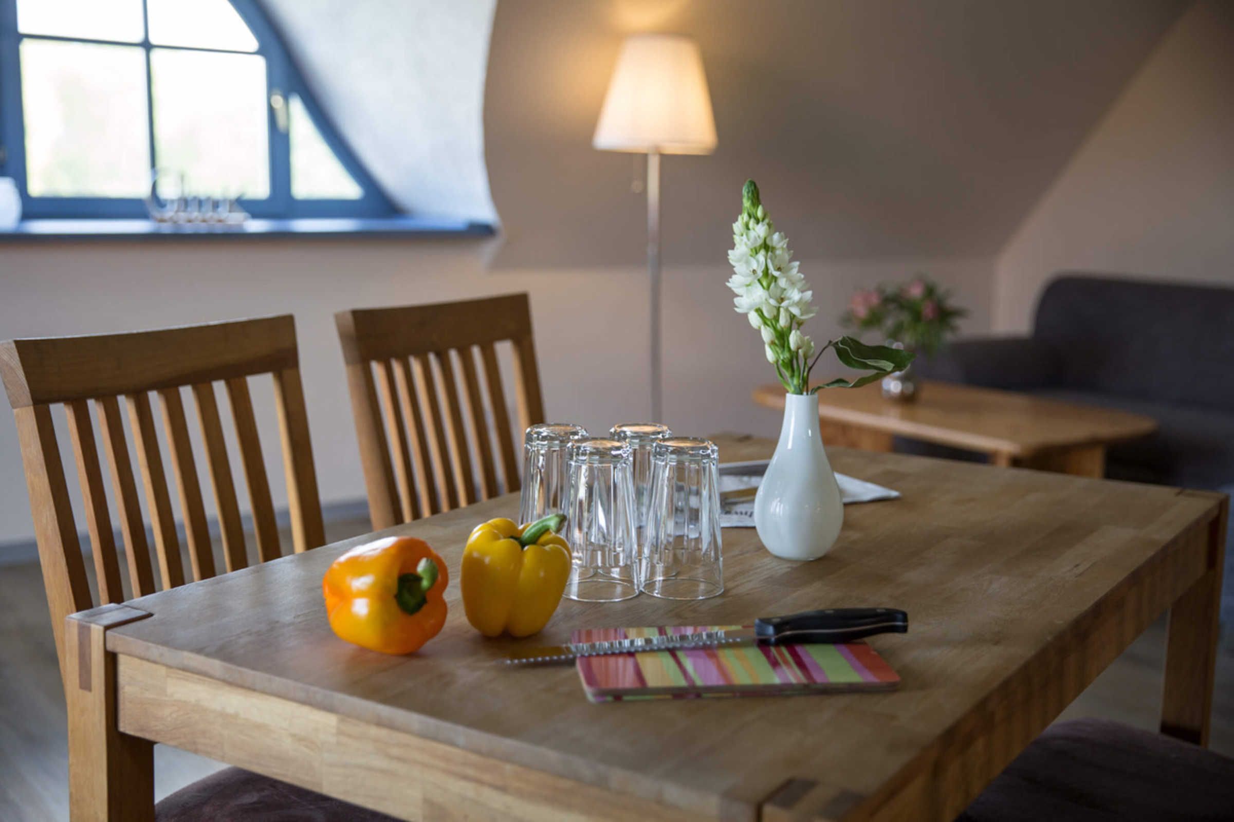 Una mesa de comedor de madera con sillas, un pimiento amarillo, un jarrón blanco con flores y un bloc de notas.