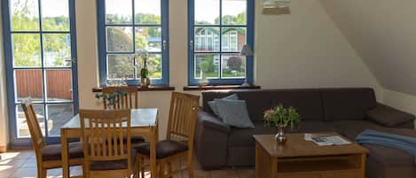 A living room with a sofa, a coffee table, a dining table with chairs, and large windows with blue frames.