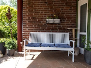 Un banco blanco con cojines azules en un patio con baldosas, una pared de ladrillo y plantas en macetas.
