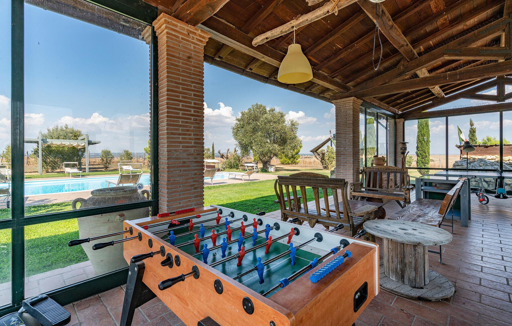 An indoor recreational area with a foosball table, a pool table, and a view of a swimming pool and greenery outside.
