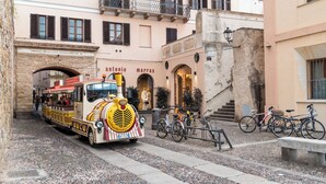 Un train d’époque dans une rue étroite, un bâtiment portant l’enseigne « Antonio Marras », et des vélos garés à proximité.