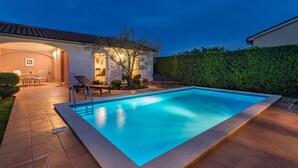 Una piscina con plataforma de baldosas, un porche cubierto con asientos y una siega al fondo.
