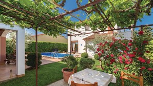 Un jardín privado con piscina, un pérgola cubierta de plantas trepadoras, una mesa con sillas y un rosal.