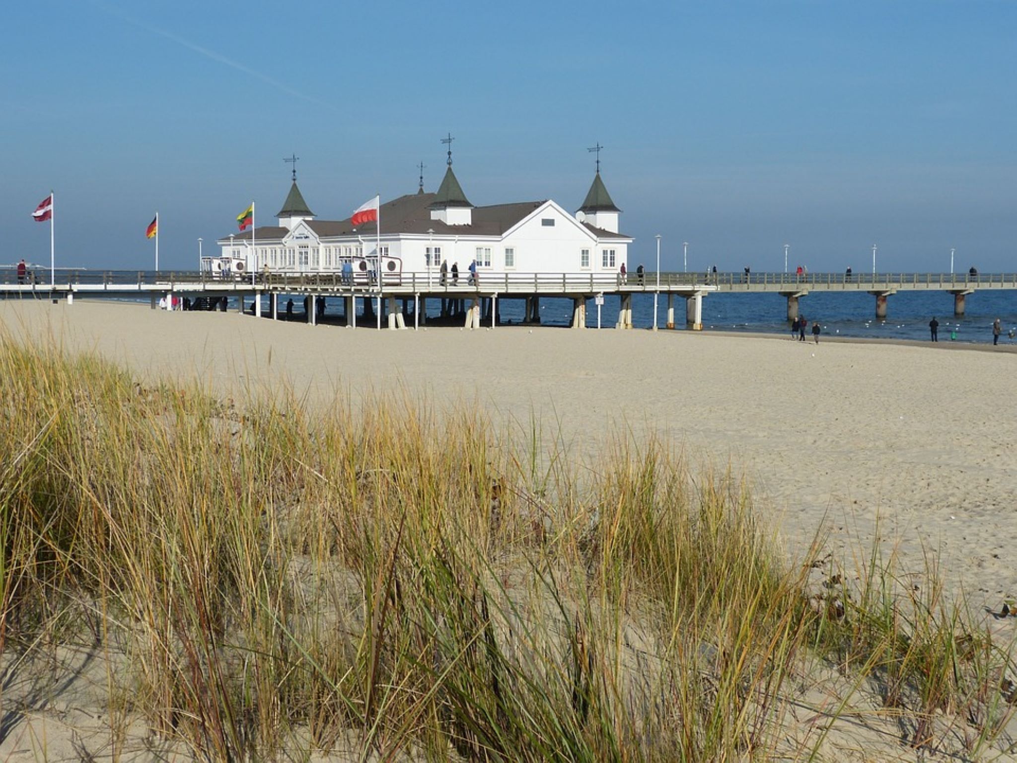 Ein weißes Gebäude mit mehreren Türmen auf Stelzen, eine Anlegestelle, die sich ins Wasser erstreckt, und Fahnen an Masten.
