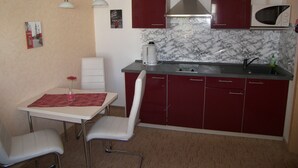 A compact kitchen with a sink, stove, and microwave. There is a dining table with two chairs and a vase with a flower. The walls are painted in a light color, and there are two pendant lights above the table. - Apartment 01 - Apartments Oechsner (Heringsdorf)