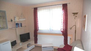 A small, well-lit living room with a television, a white sofa, a bookshelf, and a window with red curtains. - Apartment 01 - Apartments Oechsner (Heringsdorf)