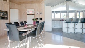 Moderni ruokailutila, jossa on puinen katto, pitkä ruokapöytä tuoleineen sekä keittiöalue, jossa on baarituoleja.