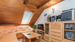 Un dormitorio tipo loft acogedor con techo inclinado, techo de madera, un ventanal grande, un sofá, una mesita de centro, un televisor, un aparador con puertas de vidrio y una impresora encima.