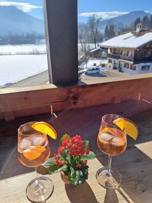 Two glasses of a beverage with orange slices on a wooden table, a small potted plant with red flowers, and a view of a snowy landscape with buildings in the background. - Blauberg vacation apartment - Nagel vacation apartments, Kreuth-Enterbach (Kreuth)