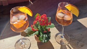 Dos vasos de una bebida con rodajas de naranja sobre una mesa de madera, una pequeña maceta con flores rojas y una vista de un paisaje nevado con edificios al fondo.
