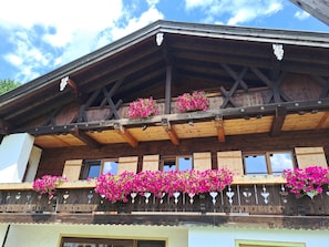 Un chalet en bois doté de balcons ornés de fleurs roses, de poutres apparentes et surplombant un ciel dégagé.