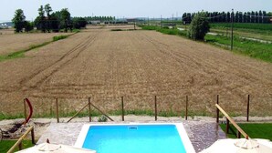 A pool area with lounge chairs and umbrellas, surrounded by a stone deck, overlooking a cultivated field. - Studio with air conditioning and pool (Ariano nel Polesine)