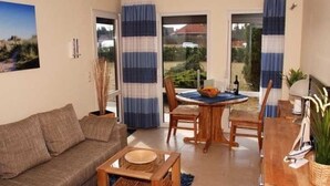 A living room with a sofa, coffee table, and dining table with chairs. There is a window with curtains, a plant, and a small sailboat decoration. - Tern (House 167) - FH 167 (Mönkebude)