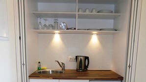 A compact kitchen with a sink, a kettle, and a shelf above the counter holding glassware and plates. - Double room 4 - PENSION Delia Will in Ahlbeck (Heringsdorf)
