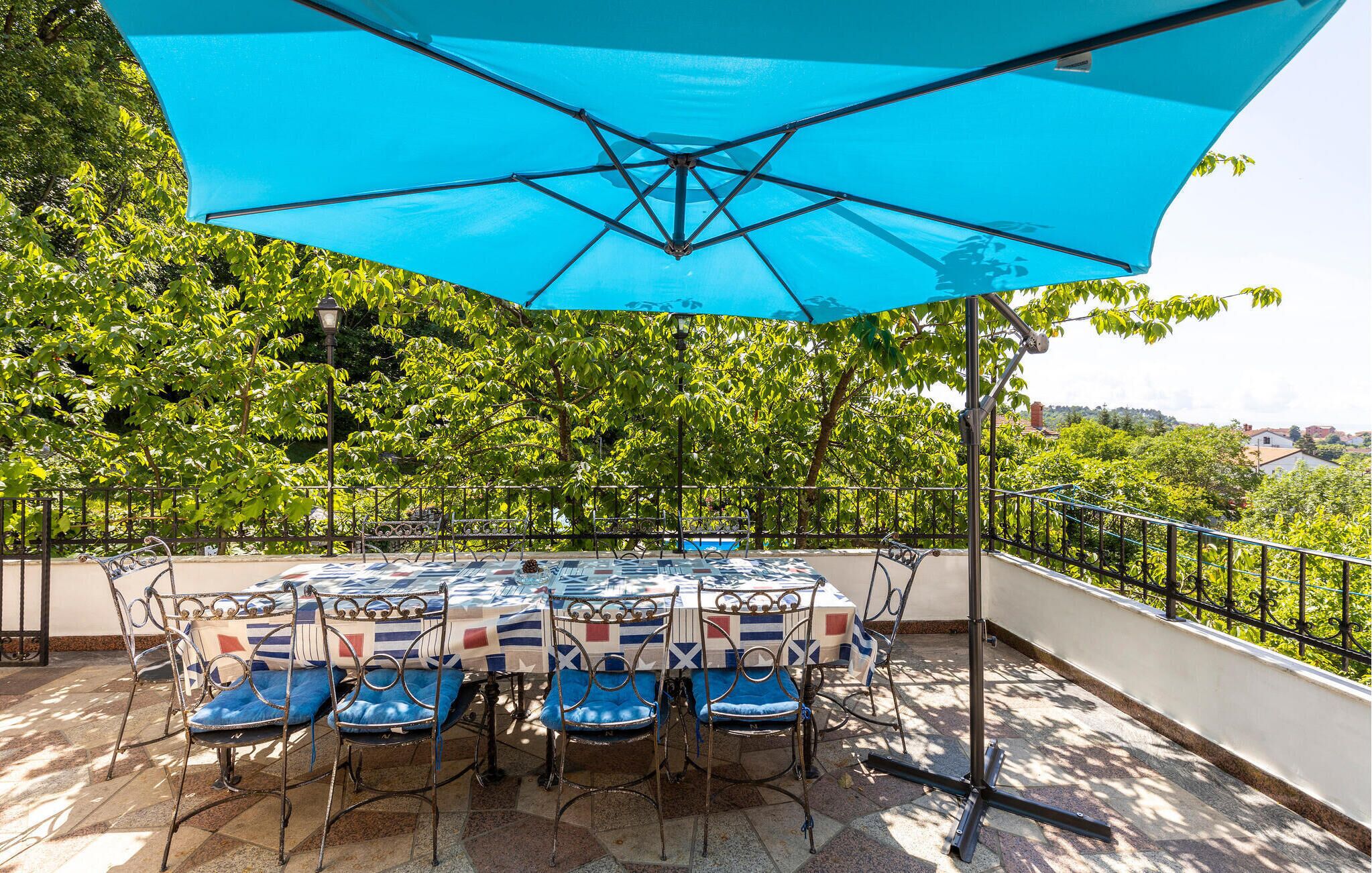 Um pátio externo com uma mesa posta para seis pessoas, rodeado por cadeiras de metal com almofadas azuis, sob um grande guarda-sol azul.