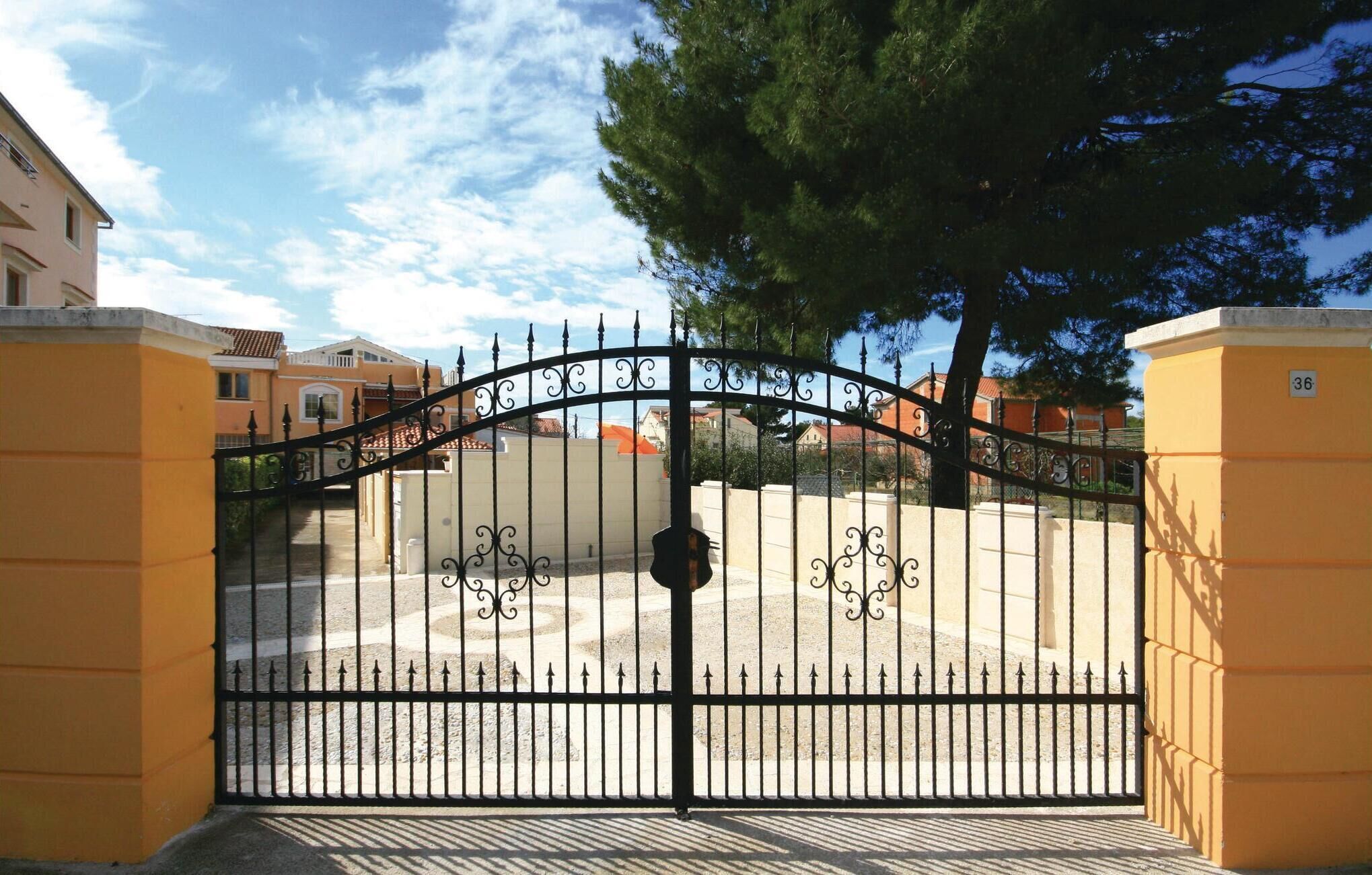 A gated entrance with a wrought iron gate, a paved driveway, and a building with a yellow facade in the background.