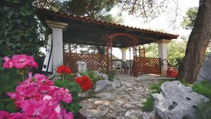 Un chemin de pierres mène à un espace de repos abrité, agrémenté d’une pergola, et est entouré de gros rochers et de plantes en fleurs.