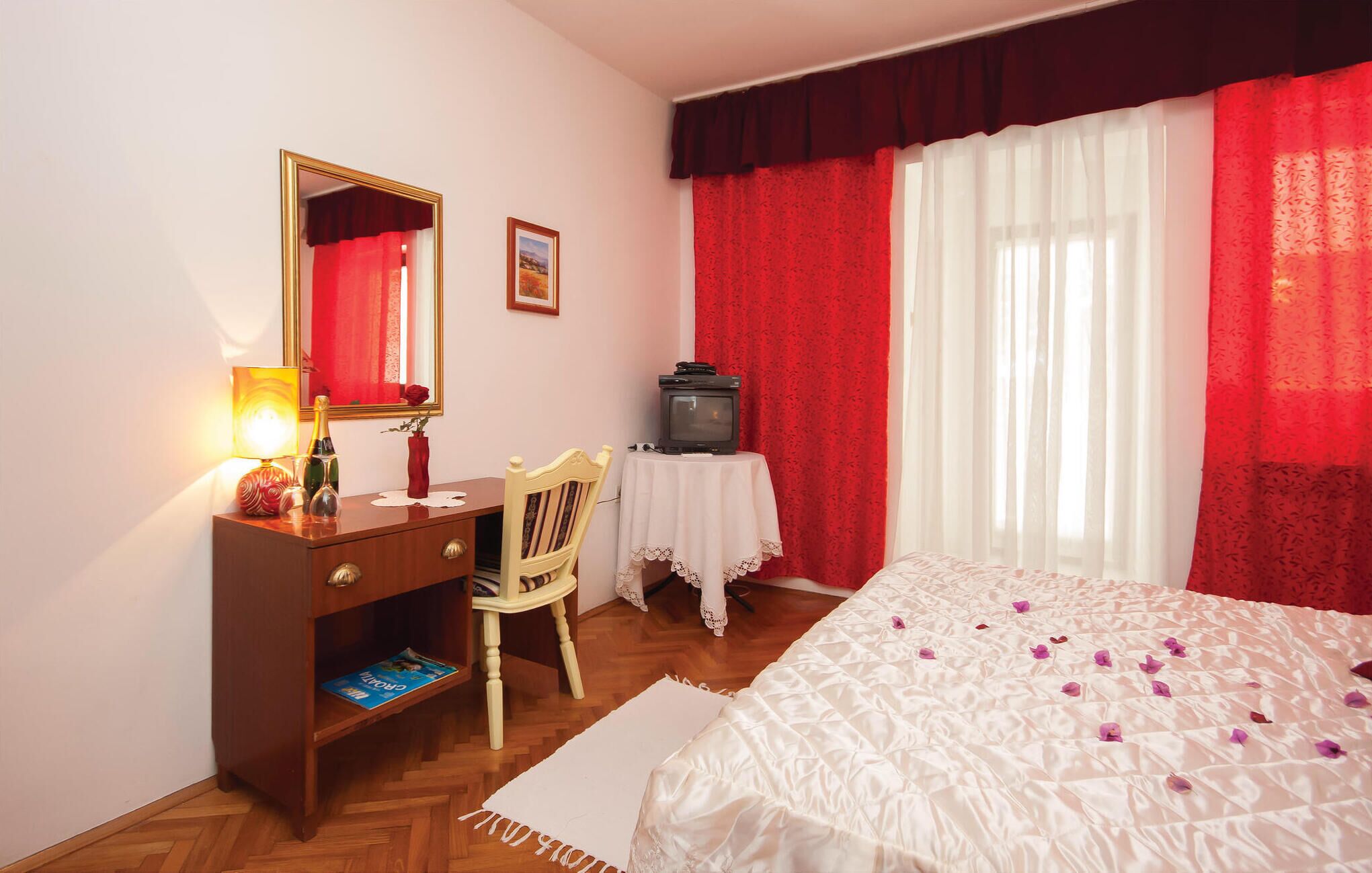 A hotel room with a bed, a desk with a chair, a television on a side table, a mirror, and a window with red curtains.